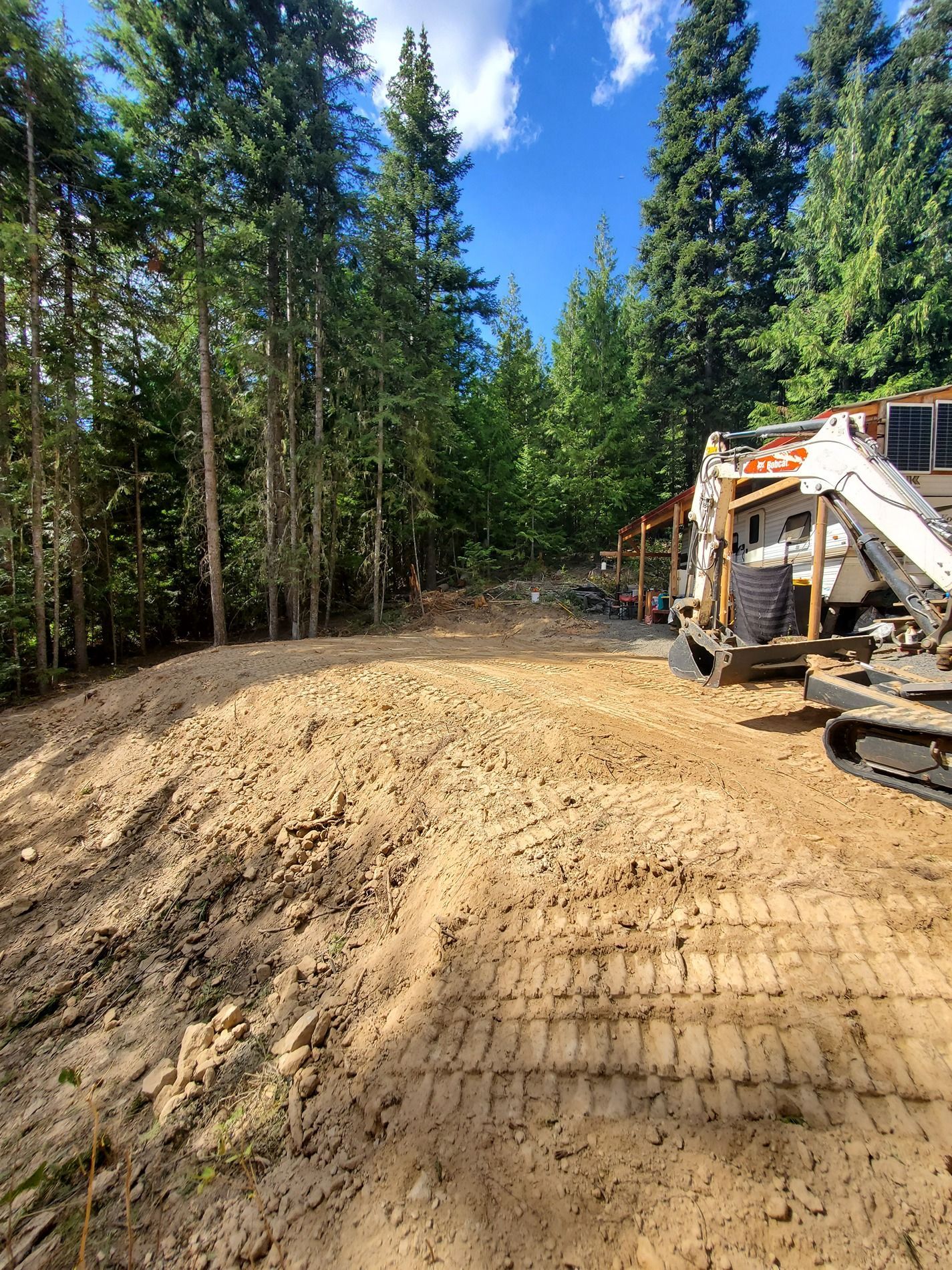 Cleared dirt area with an excavator, surrounded by trees under a blue sky.