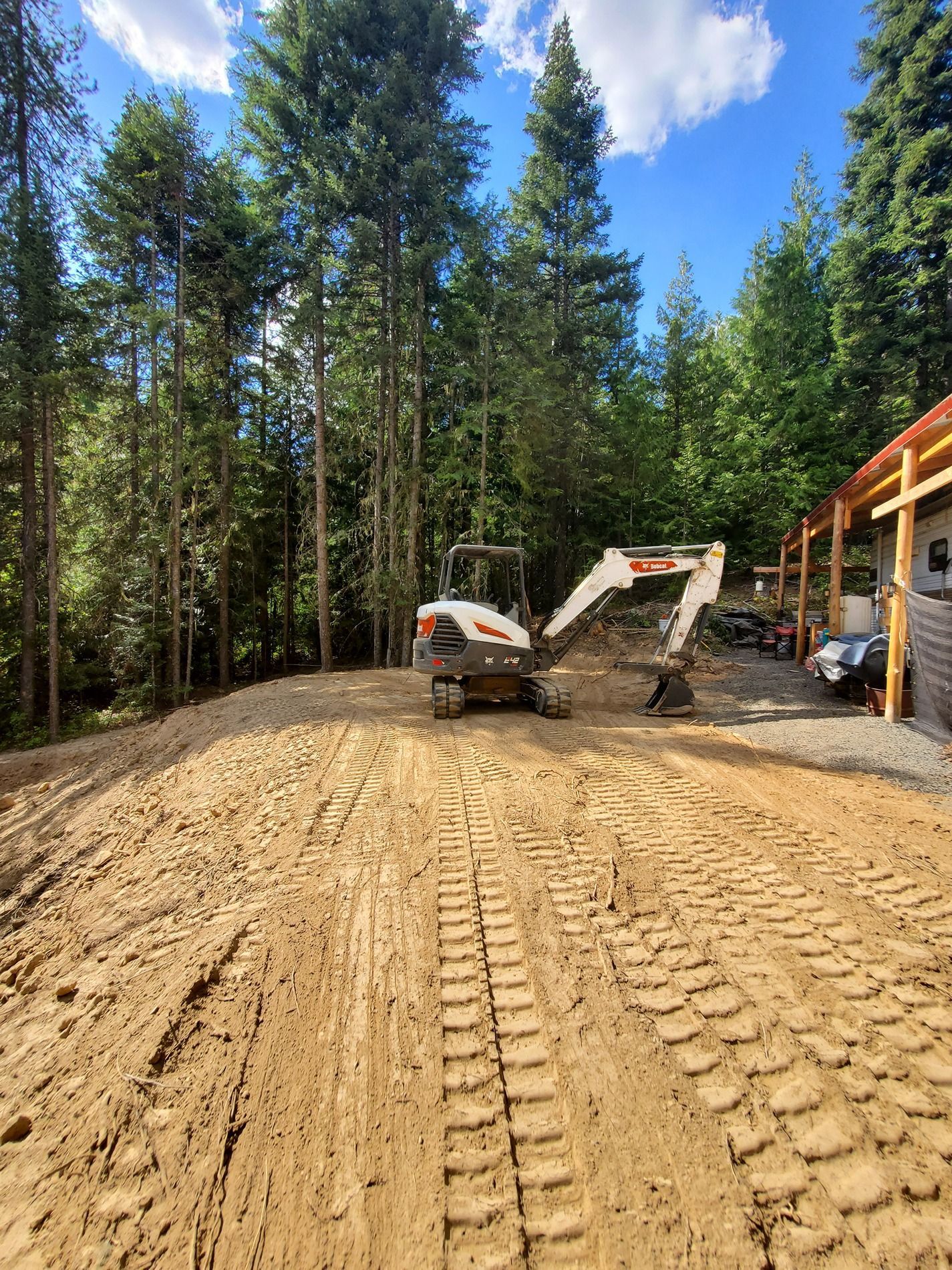Mini excavator on a graded dirt surface, surrounded by trees and a building on a sunny day.