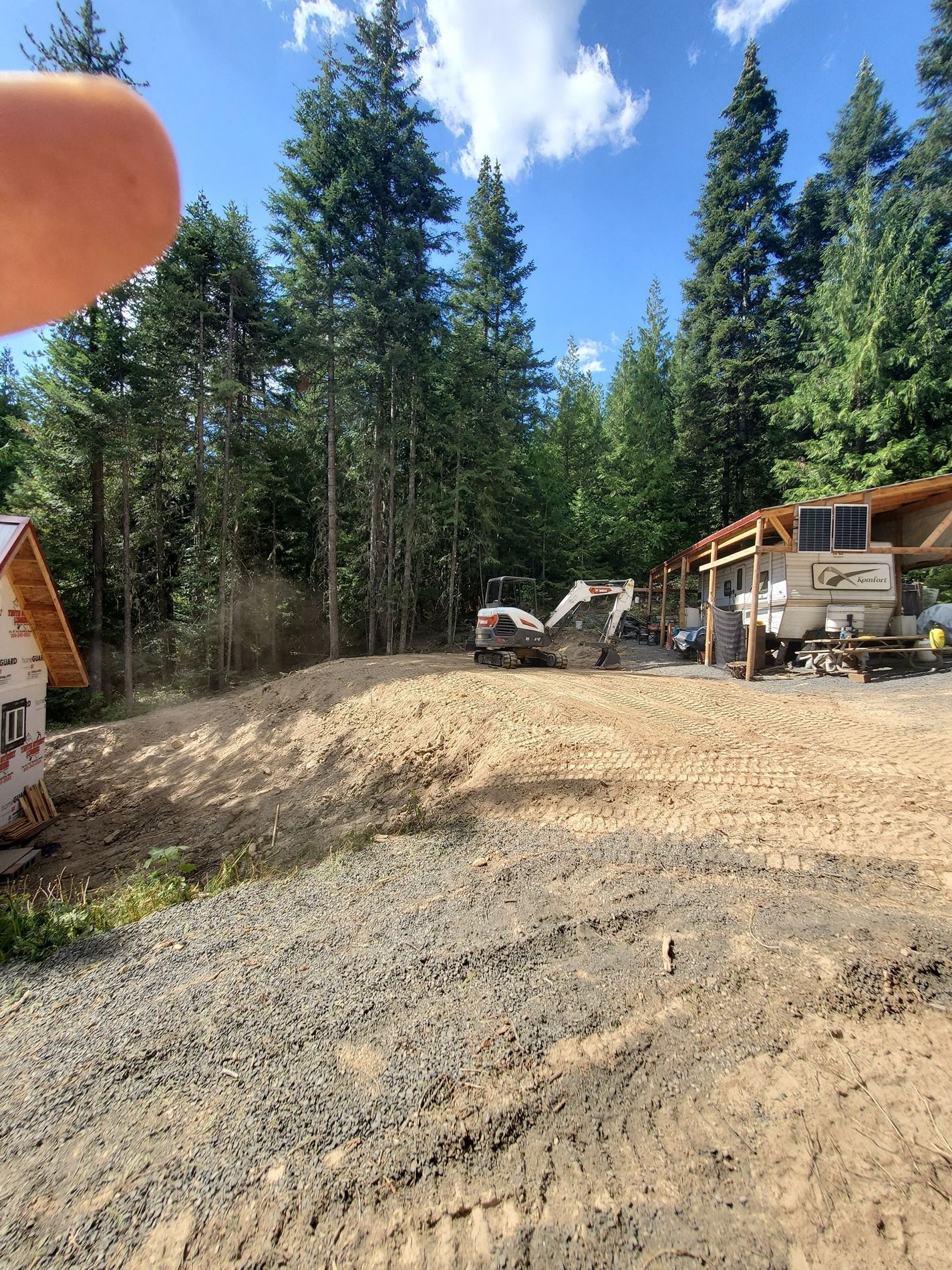 Construction site with a backhoe, dirt pile, gravel, and trees under a blue sky.