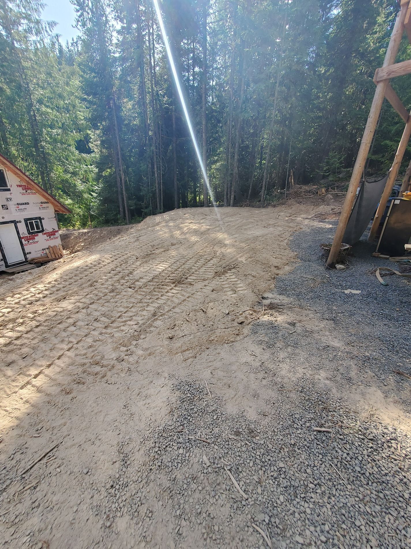 Dirt path through a construction site, leading toward a wooded area under a bright sky.