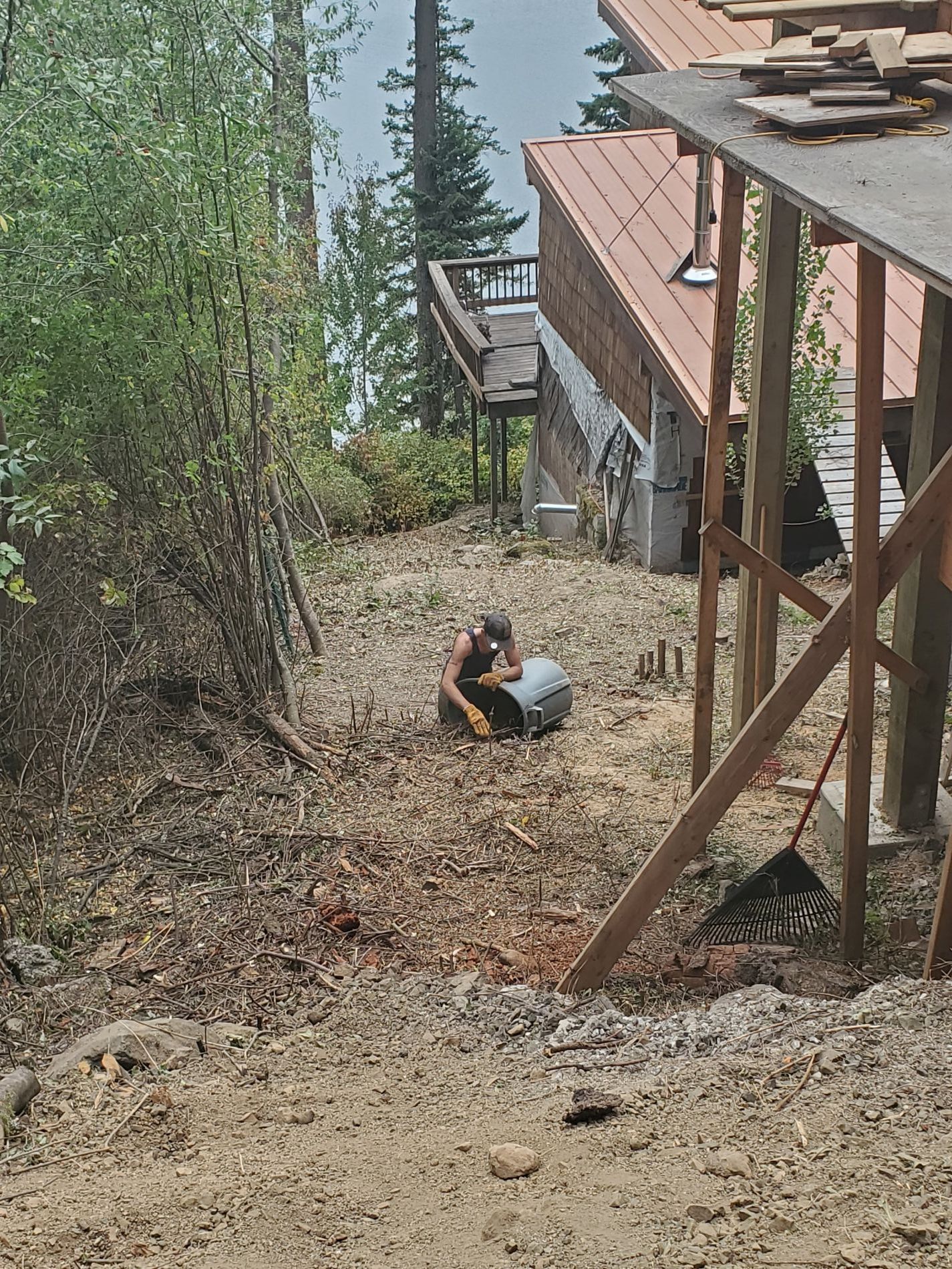 Person seated outdoors with a metal container, working on a sloped dirt path next to a building.