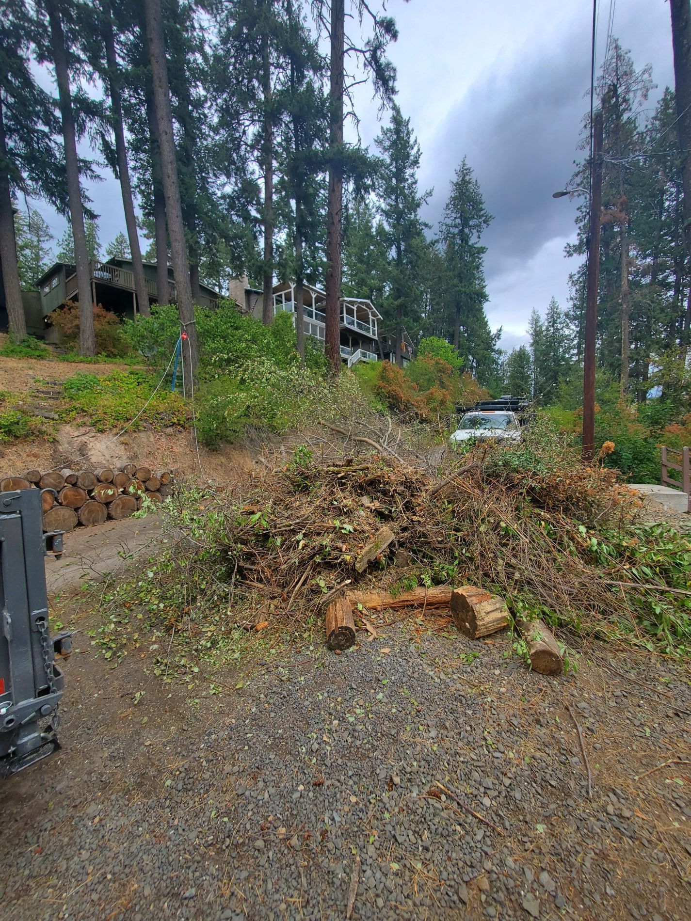 Pile of brush and logs on a gravel driveway, with a truck parked nearby and a house on a hillside in the background.