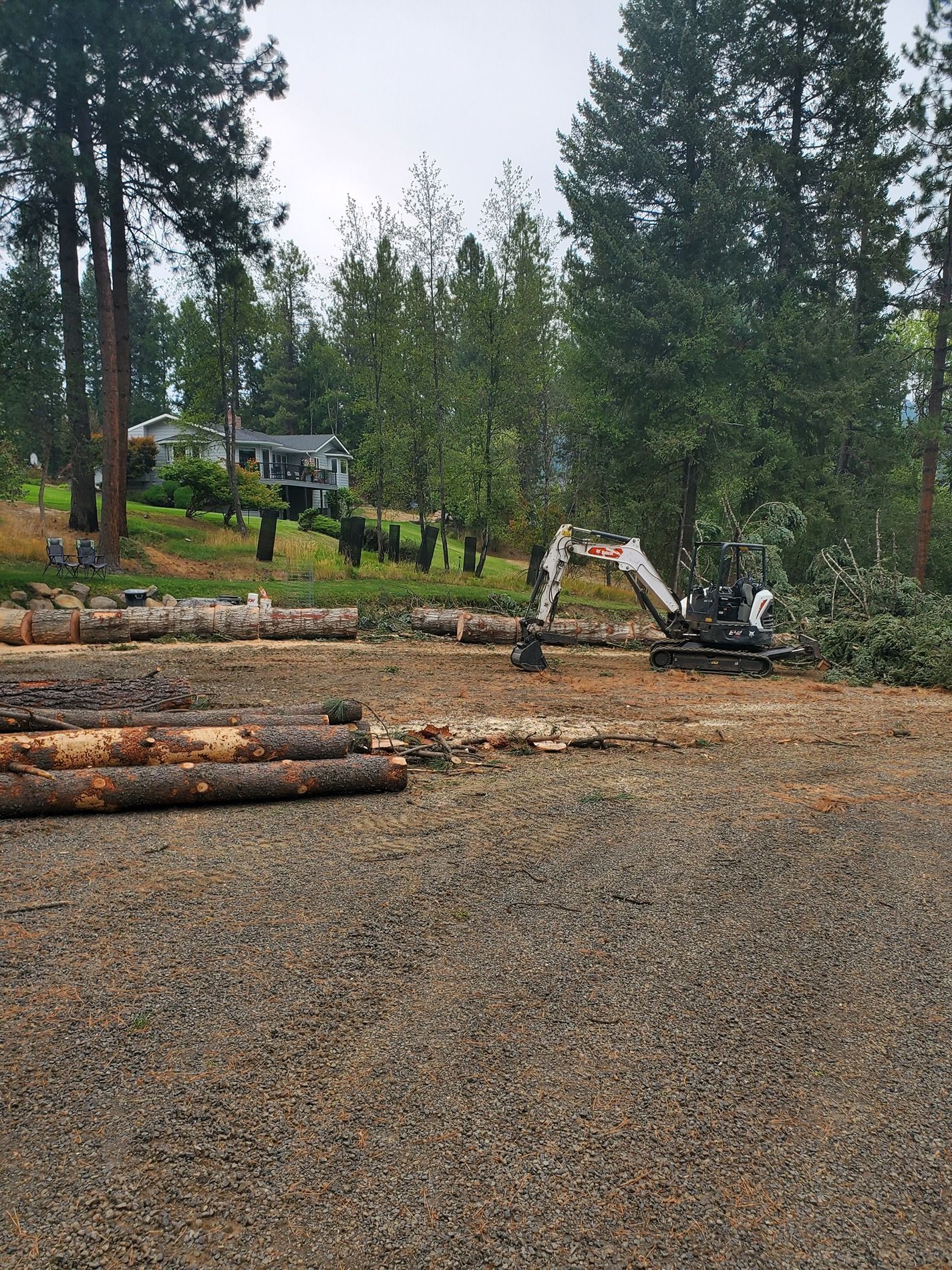 Mini excavator removing logs from a gravel clearing, with a house and trees in the background.
