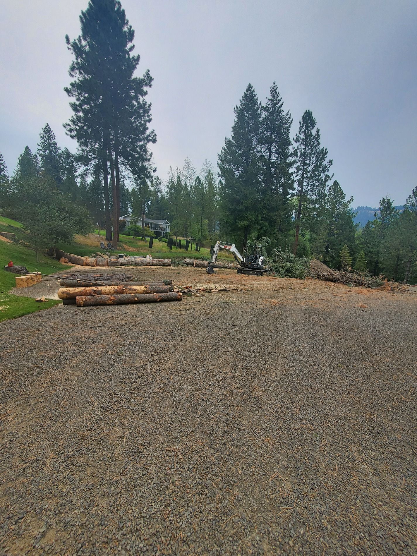 Construction site with gravel, logs, excavator, and trees. Overcast sky.