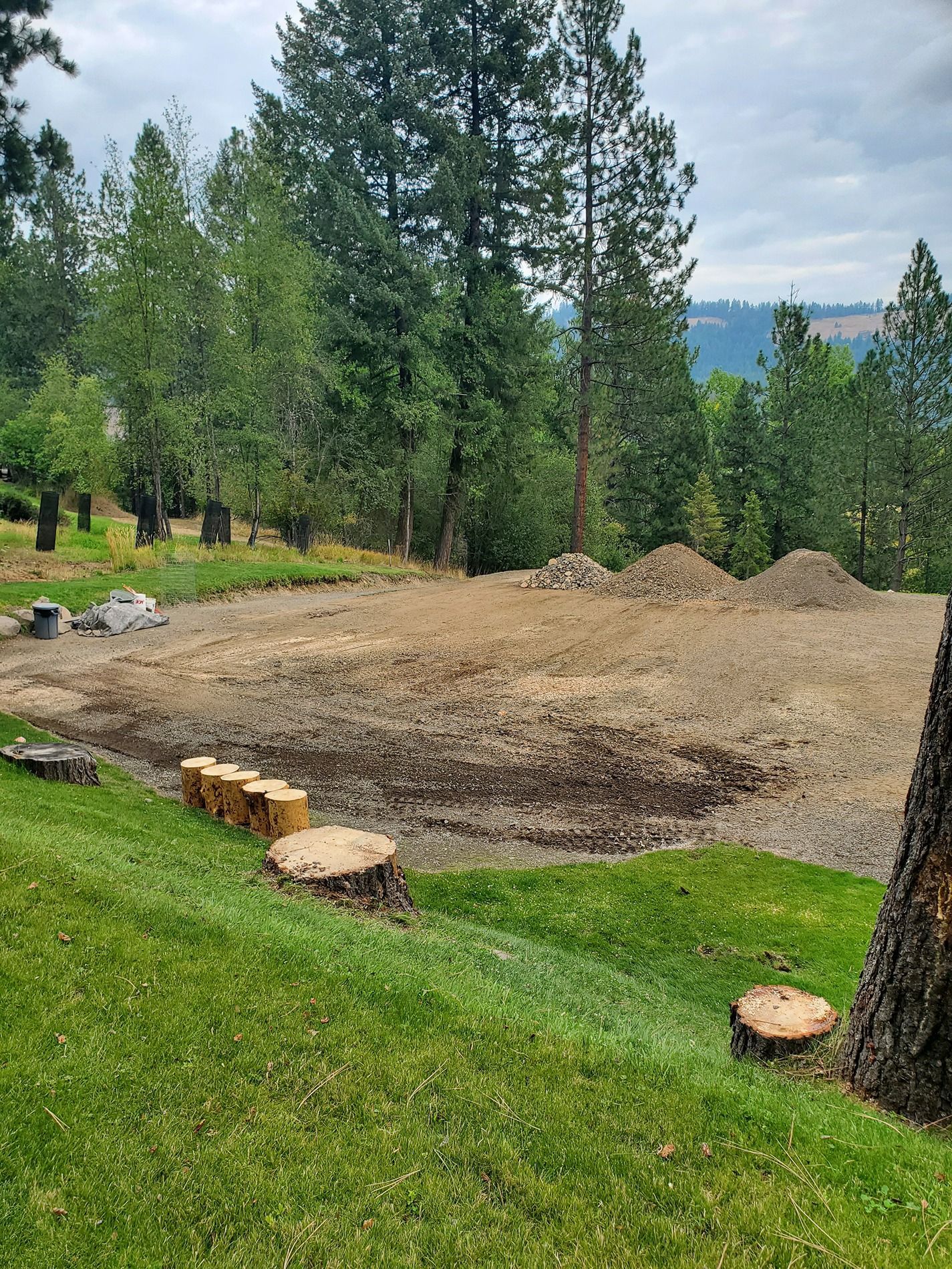Dirt lot with piles of dirt, tree stumps, and green grass against a backdrop of trees and a cloudy sky.