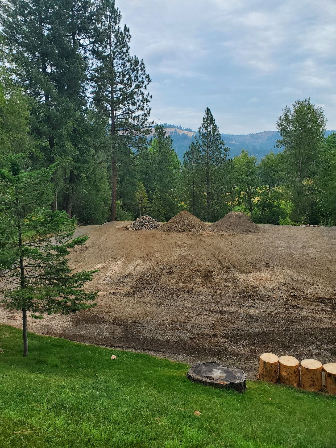 Excavated dirt piles on a leveled area, surrounded by trees and a view of distant hills. Cloudy sky.