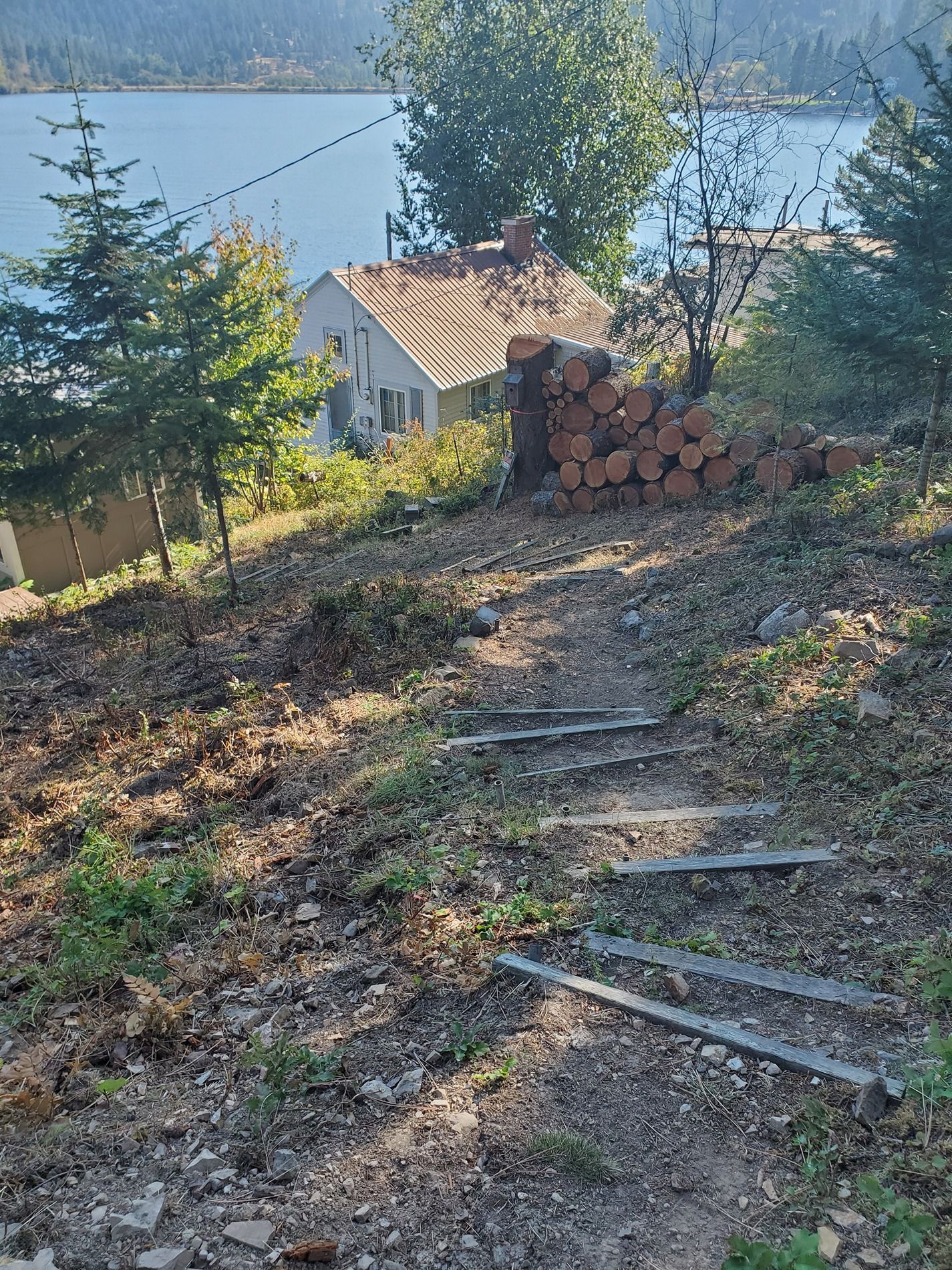 A hillside path with steps leads to a house near a lake. Firewood is stacked nearby.