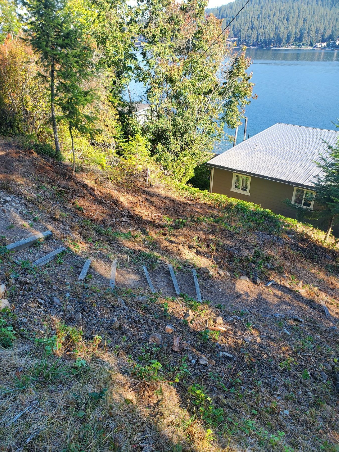 Sloped, bare ground with wooden steps leading down to a waterfront house. Trees and water in the background.