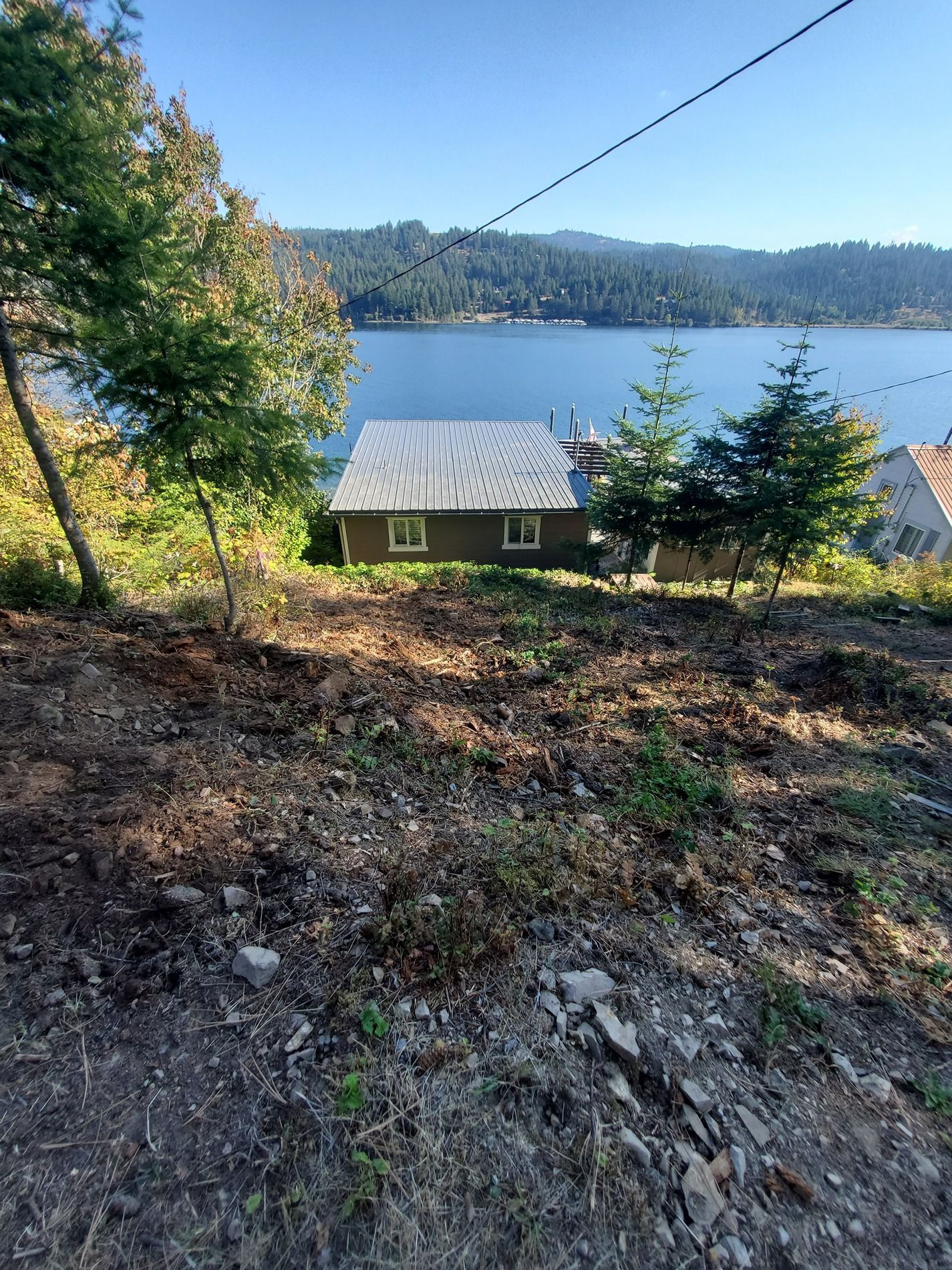 A house on a hillside overlooking a lake, with trees in the foreground and a blue sky.