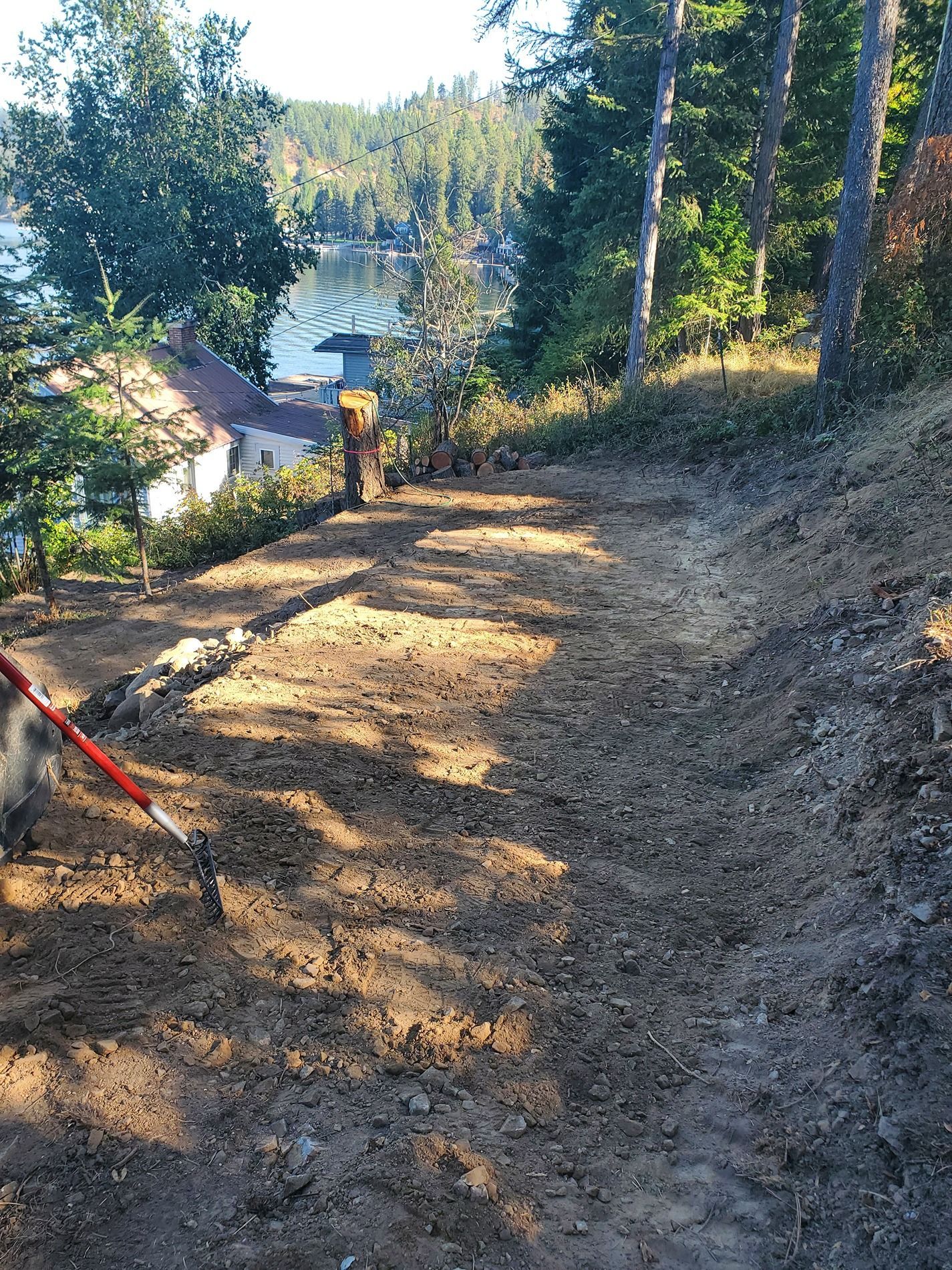 A dirt path slopes downward towards a lake, lined with trees on one side. A house is visible in the background.