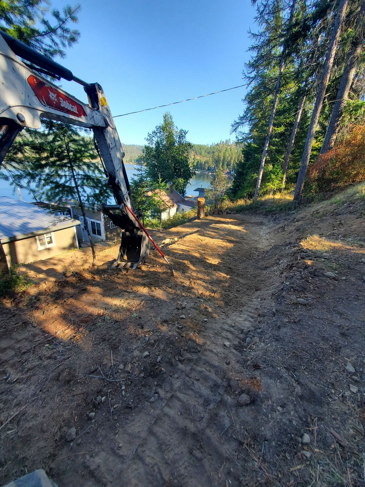 Bobcat excavator clearing a dirt path on a hillside with a lake and trees in the background.