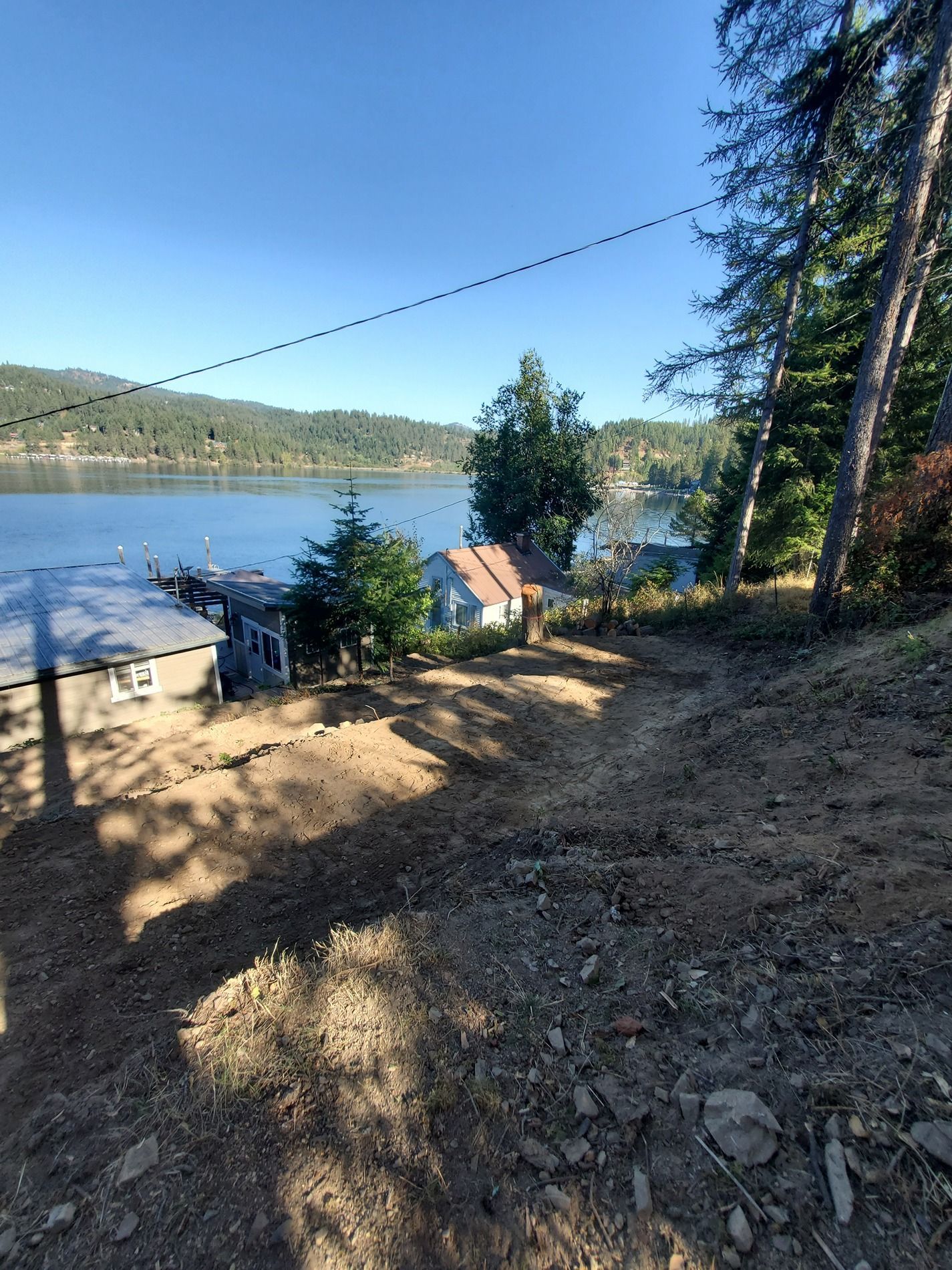 Dirt path leading down to a waterfront with houses and a dock under a blue sky.