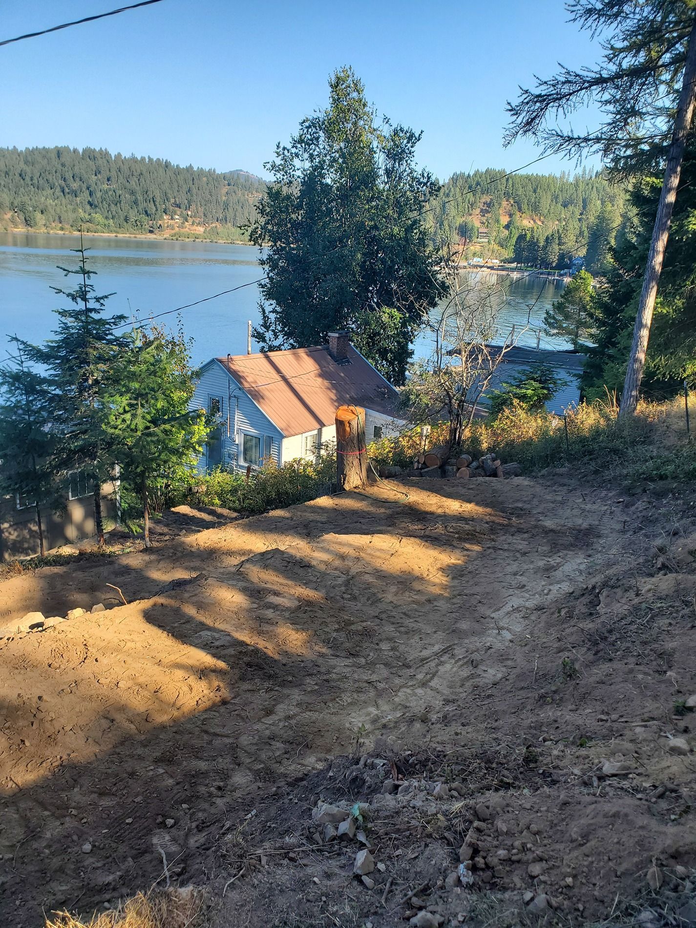 Dirt path leading down to a blue house with lake and hillside view. Sunny day.