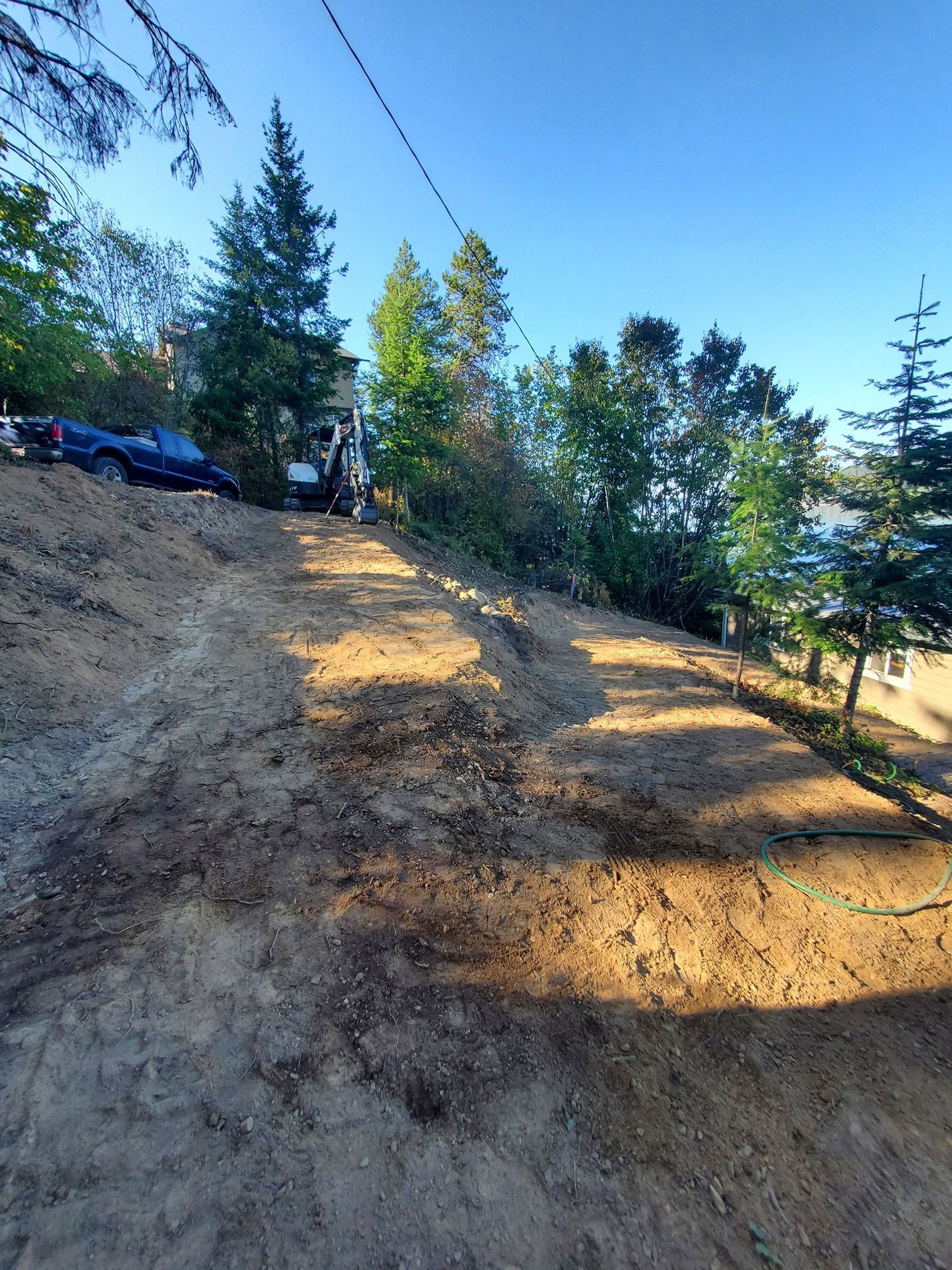 Dirt hillside with tire tracks, trees, and buildings visible in the distance under a blue sky.