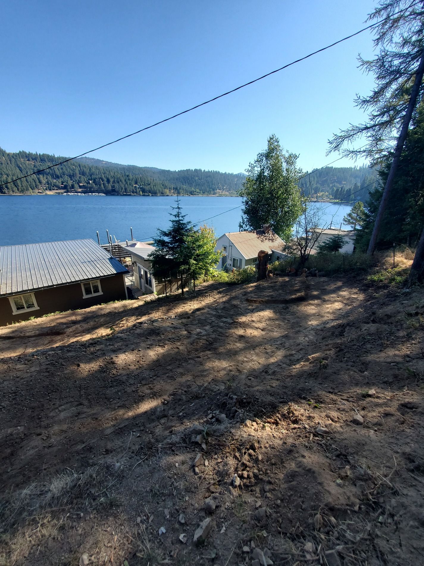 Dirt lot with lake view, houses and trees in the background. Blue sky.