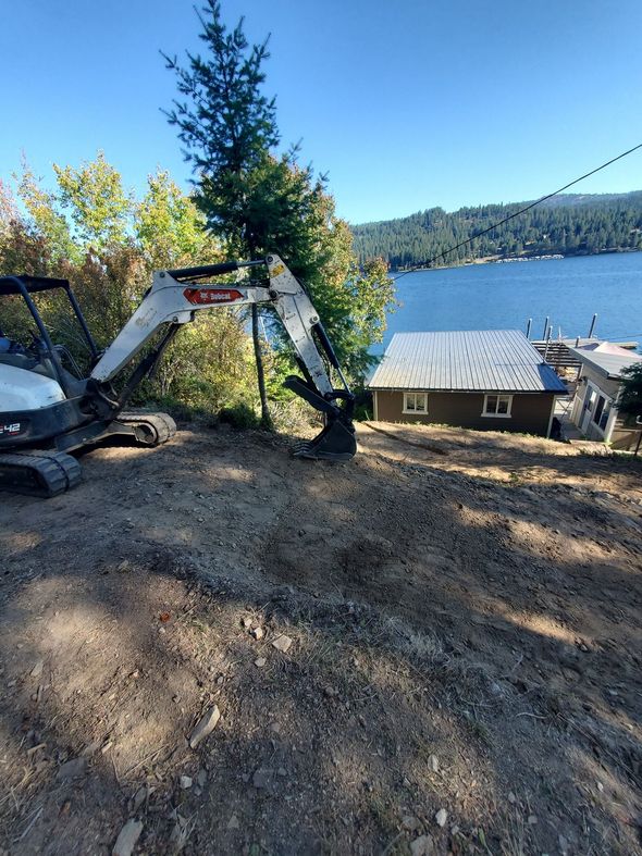 Bobcat excavator on a hillside, overlooking a lake with a small cabin. Sunny day.