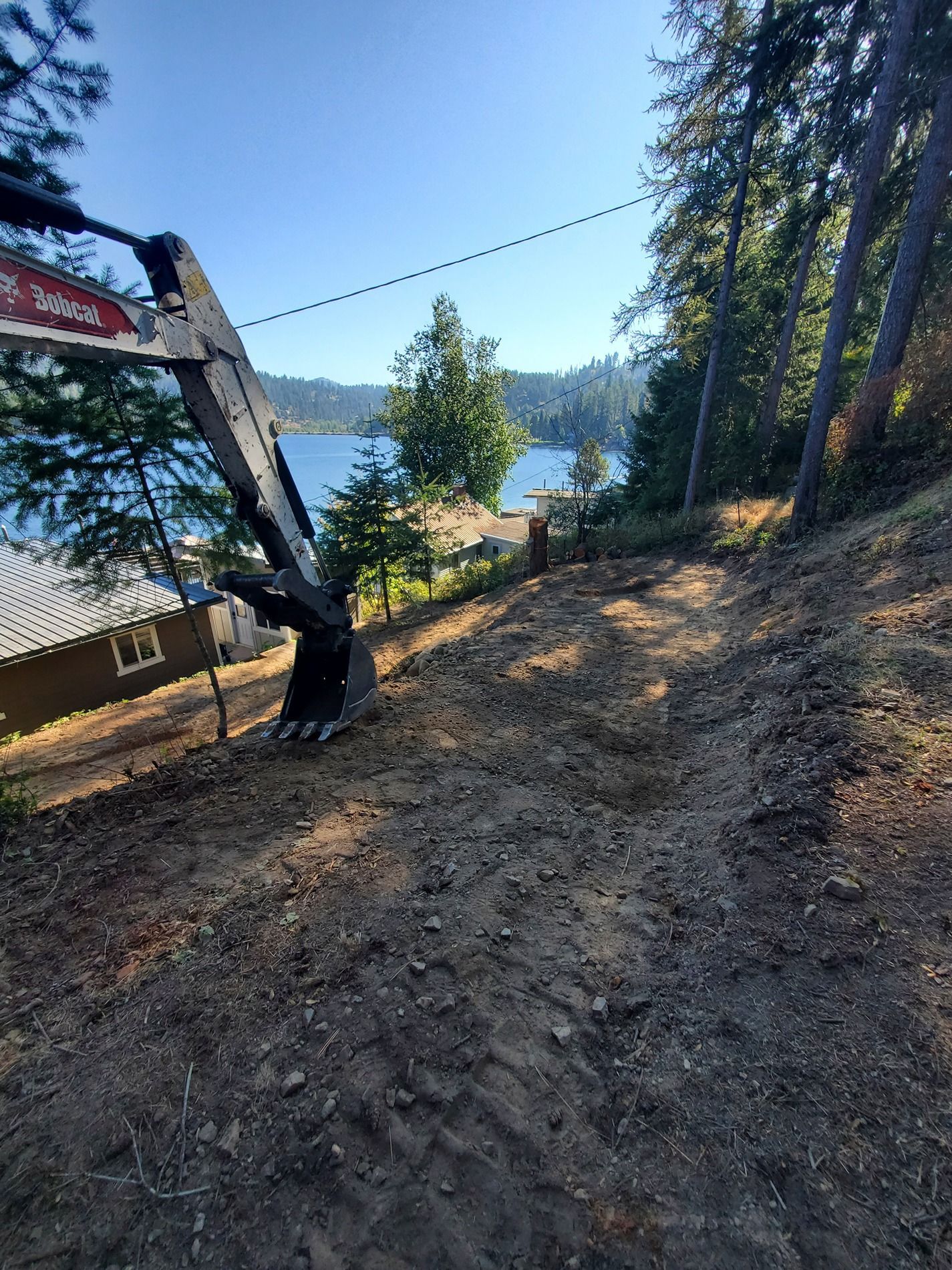 Excavator on a hillside clearing dirt. Lake and trees in the background under a blue sky.