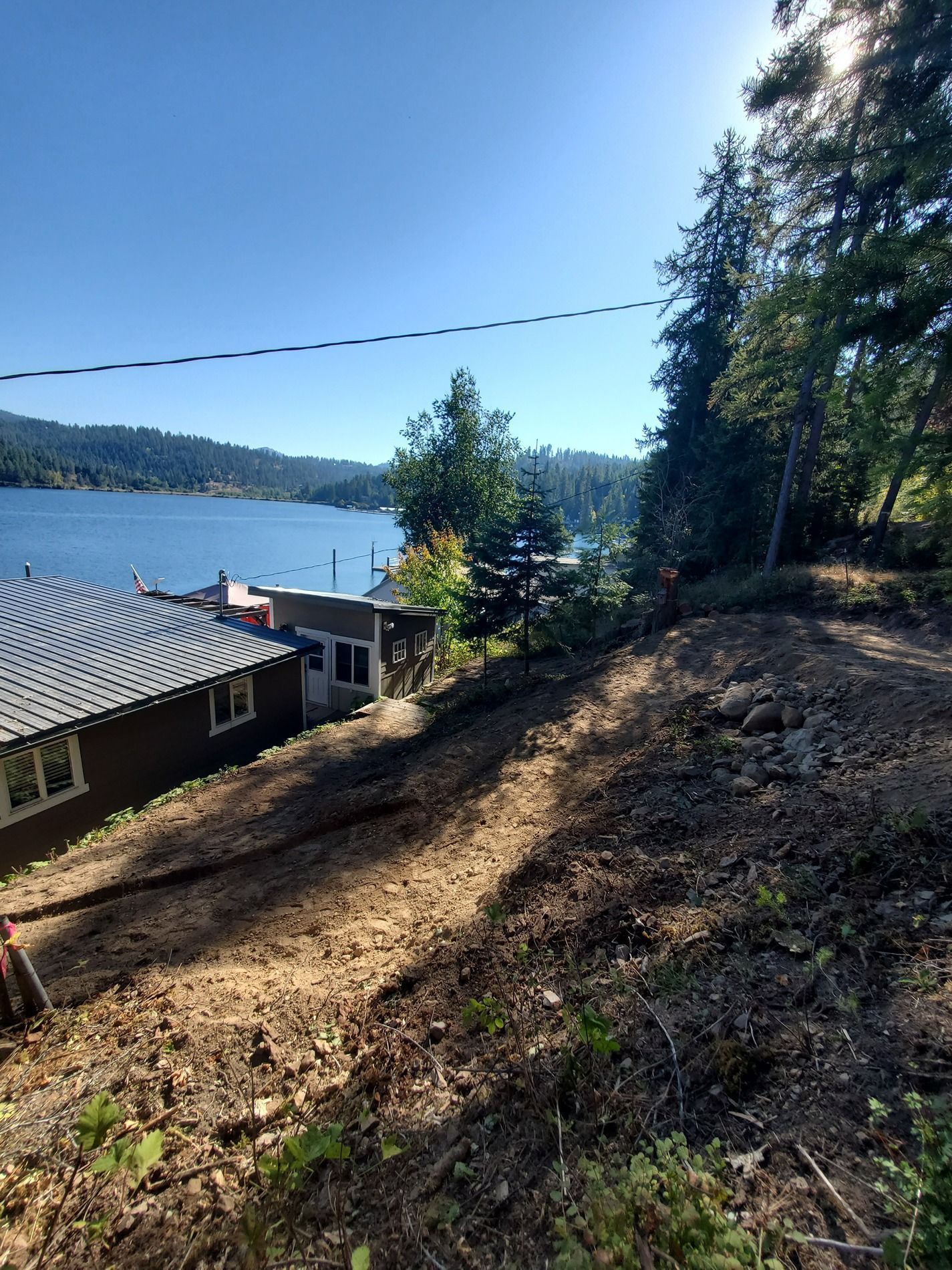 Sloped, cleared dirt area with a view of a lake and shoreline houses under a sunny sky.