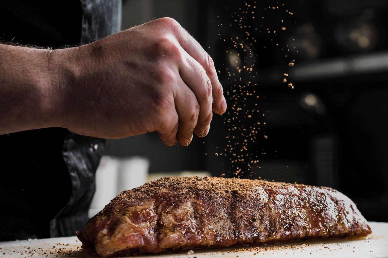 A person is sprinkling seasoning on a piece of meat on a cutting board.