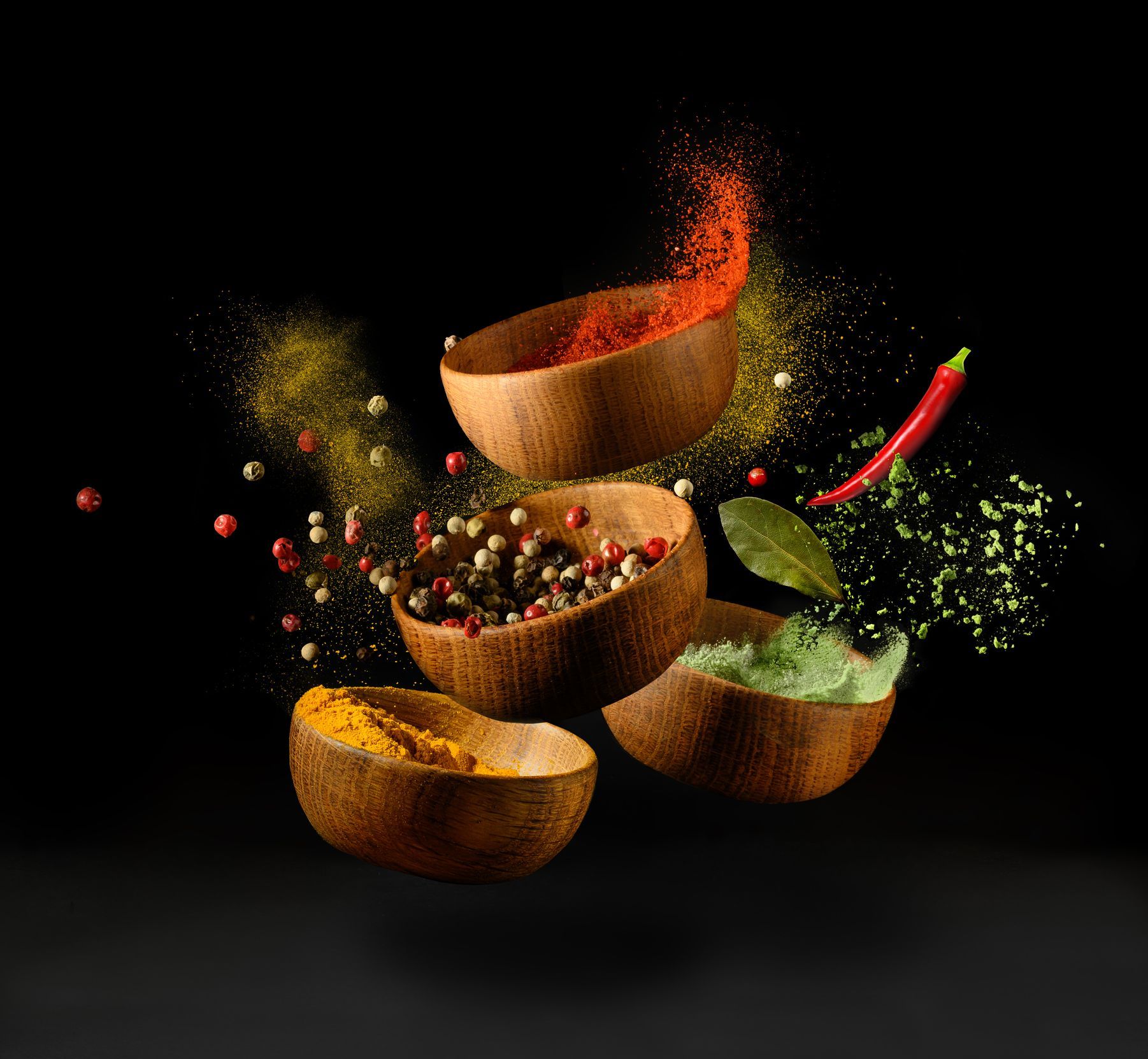 A variety of spices are falling into wooden bowls on a black background.