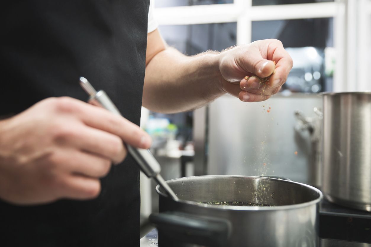 A man is adding salt to a pot of food in a kitchen.