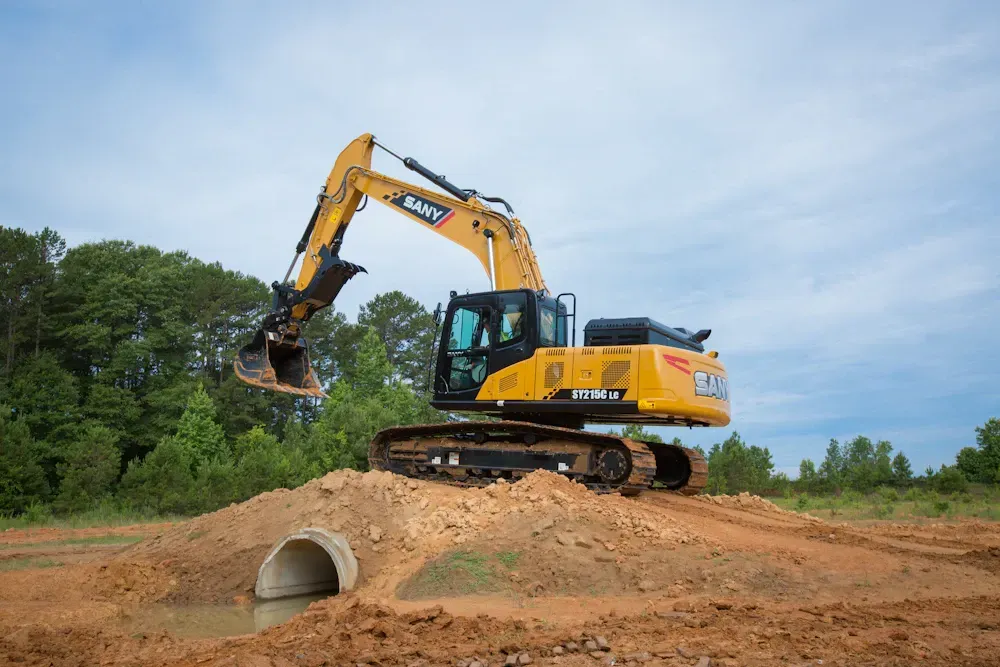 Yellow SANY excavator on a dirt mound over a culvert, in a field.