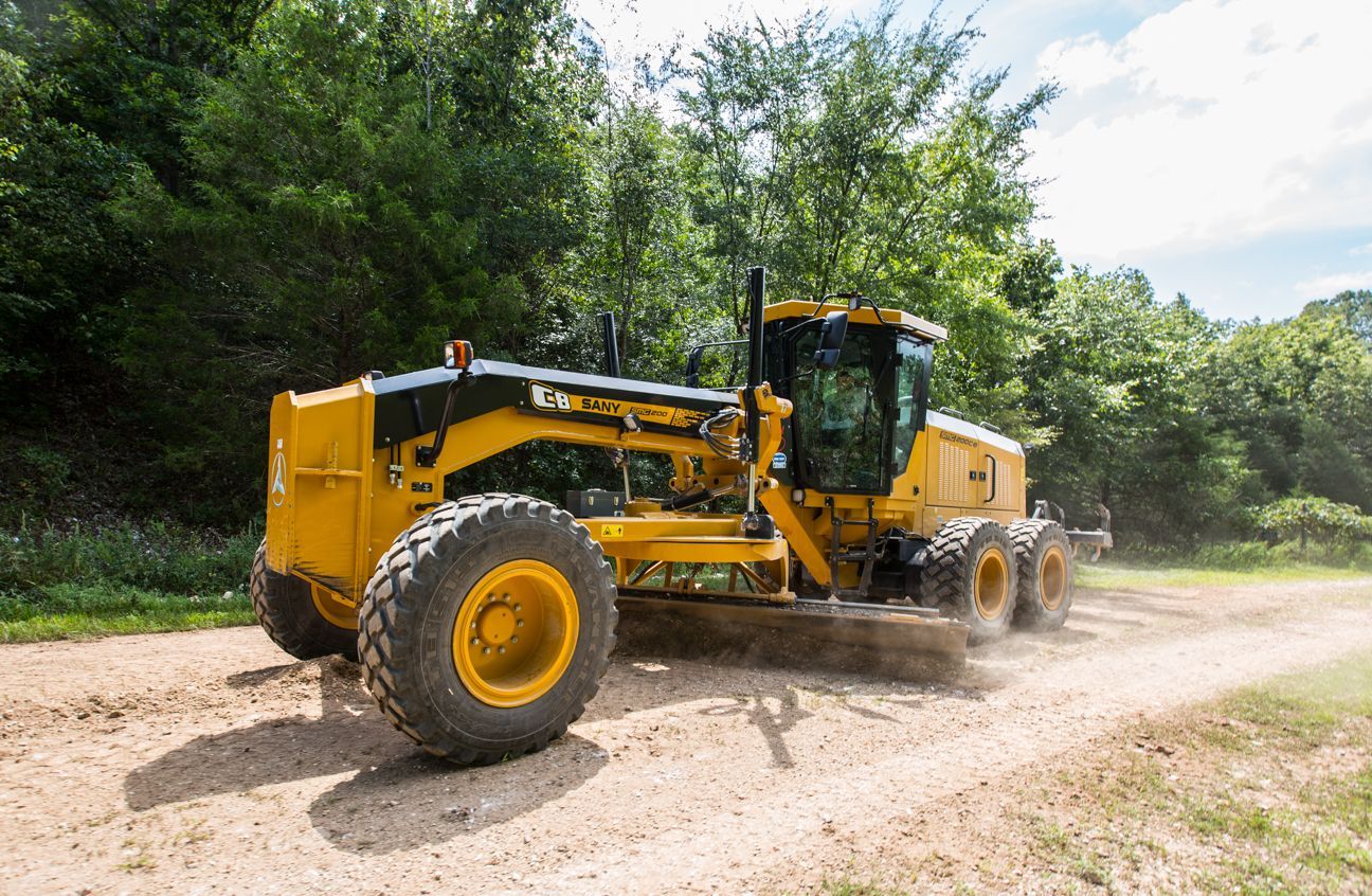 Yellow Caterpillar road grader smoothing a dirt road in a wooded area.