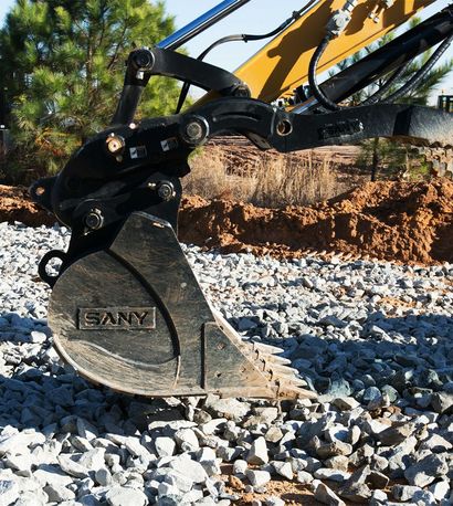 Excavator bucket, black and yellow, digging into gravel, sunny day.