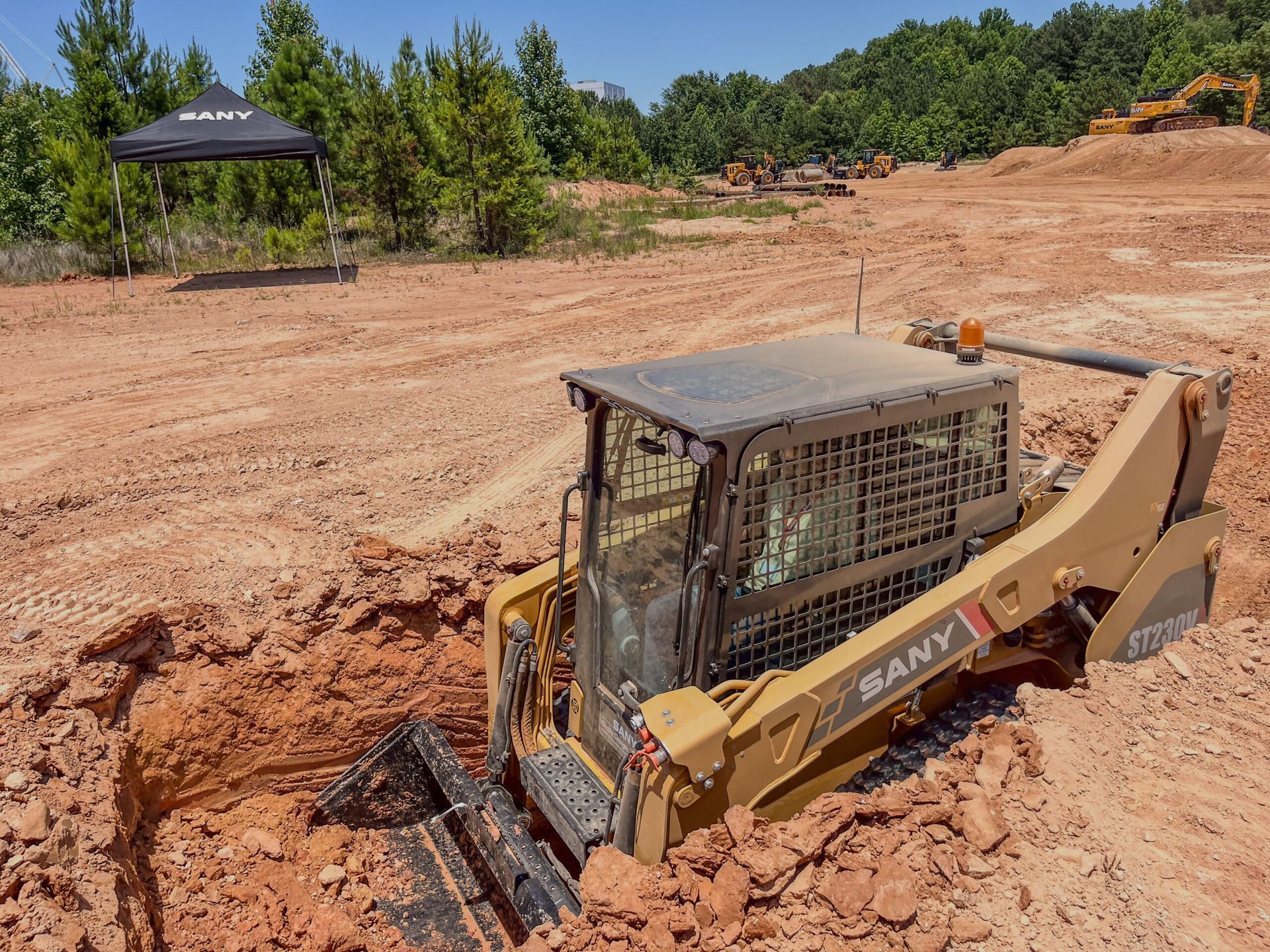 A yellow SANY skid steer loader operates in a red dirt construction site near a small, black branded canopy tent.