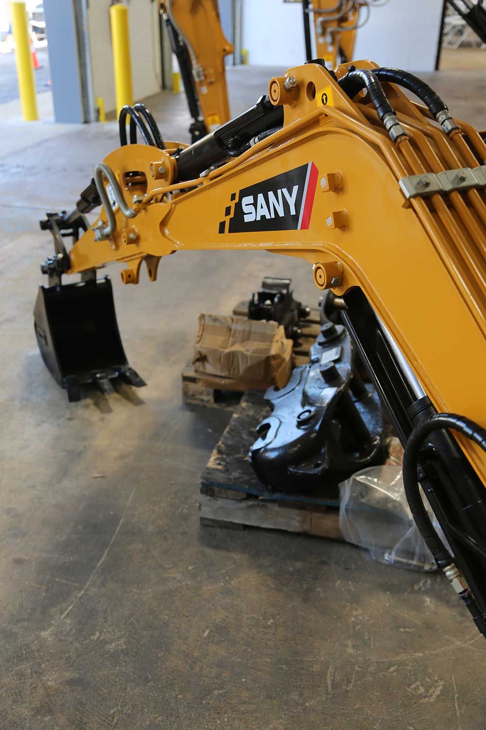 Yellow SANY excavator arm and bucket over a pallet, in an indoor space with concrete flooring.