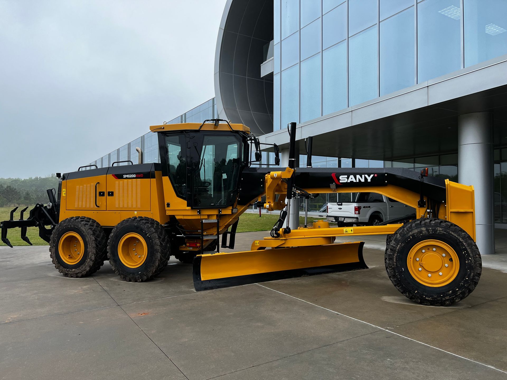 Yellow SANY motor grader parked outside a modern building with glass windows.