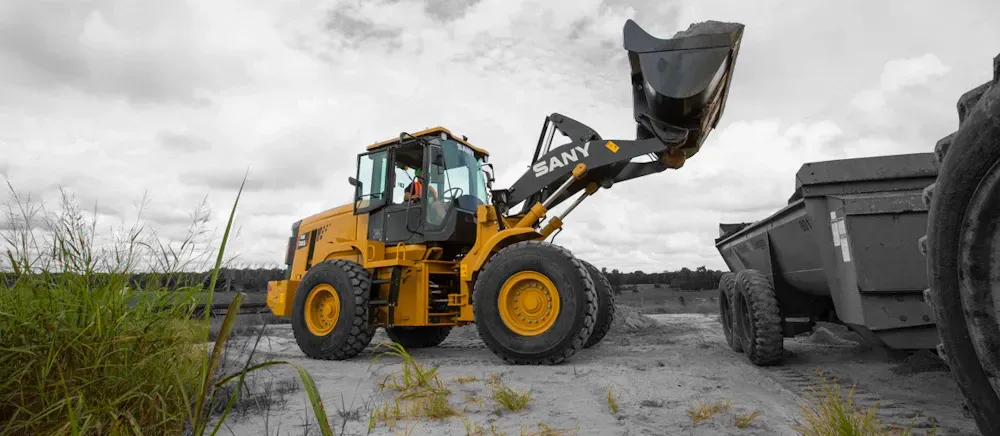 Yellow loader with bucket raised, filling black trailer, in overcast field.