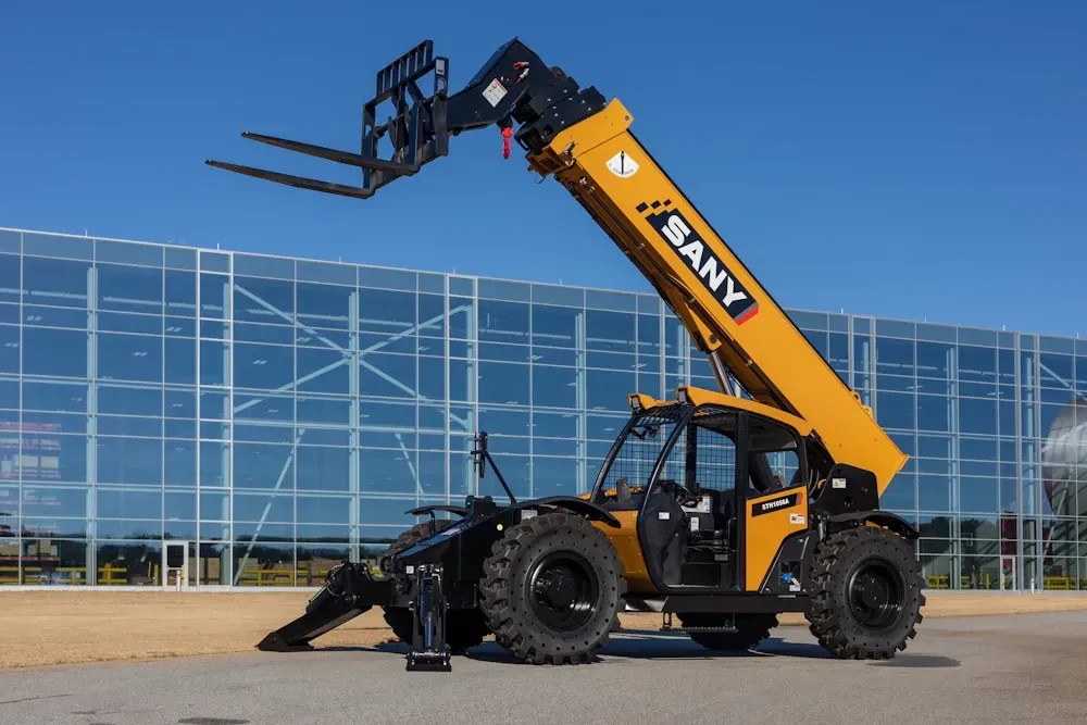 Yellow SANY telehandler with extended boom and forks, black wheels, against a glass-walled building and blue sky.