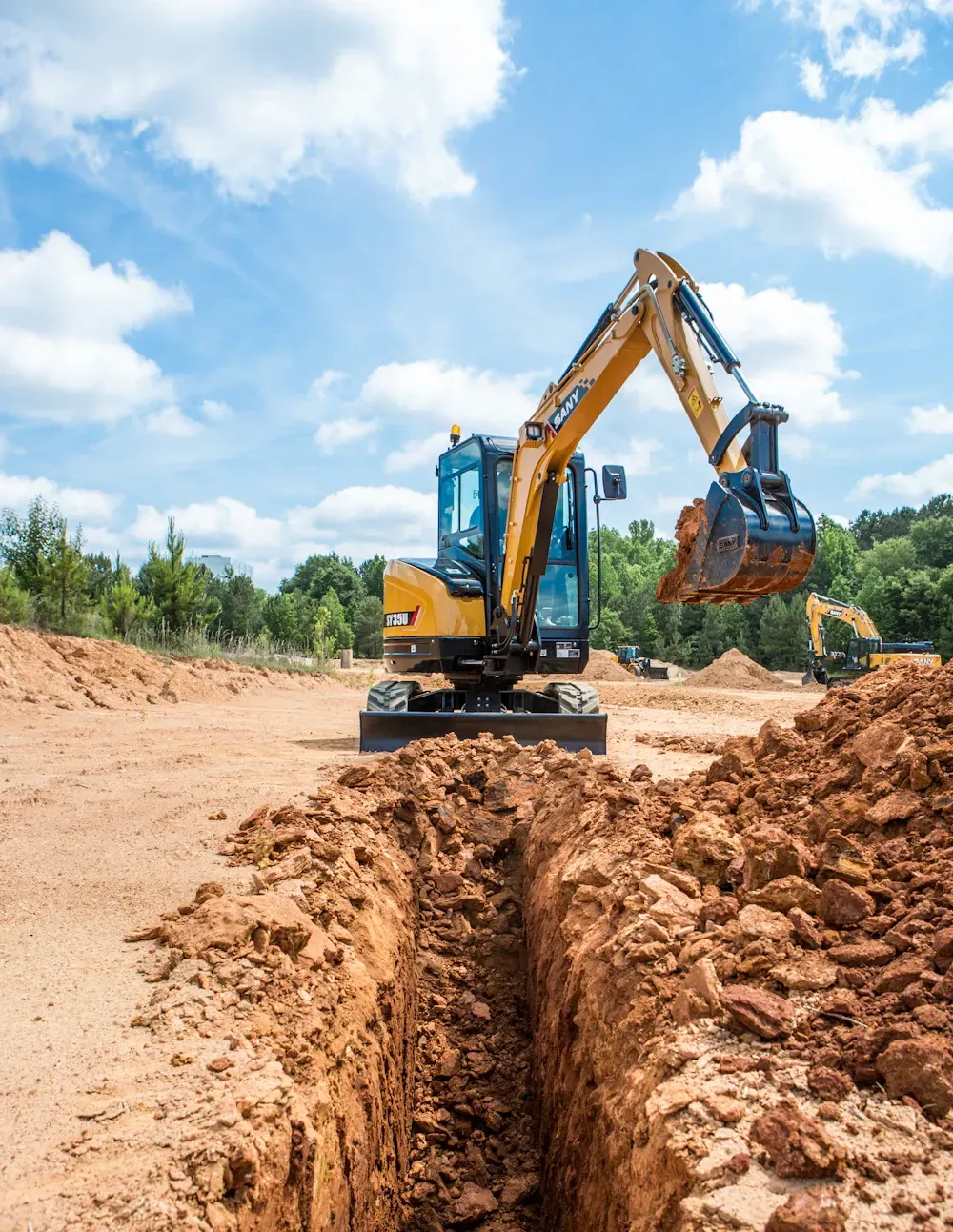 Yellow excavator digging a trench in a dirt field on a sunny day.