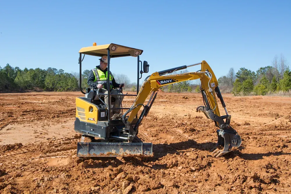 Person operating a small yellow excavator on a muddy worksite on a sunny day.