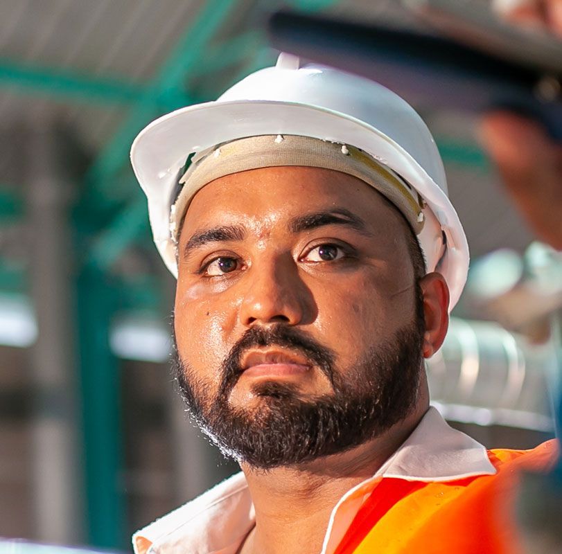 Man in hard hat, wearing orange safety vest, looking serious. Outdoors, likely construction site.