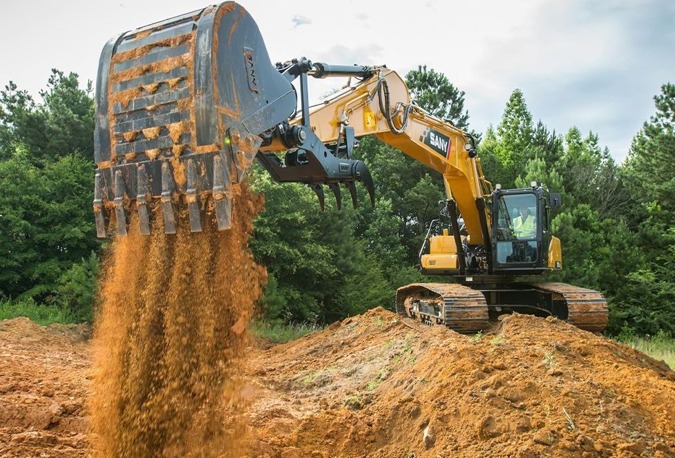 Excavator dumping red soil. Yellow machine, brown earth, green trees, overcast sky.