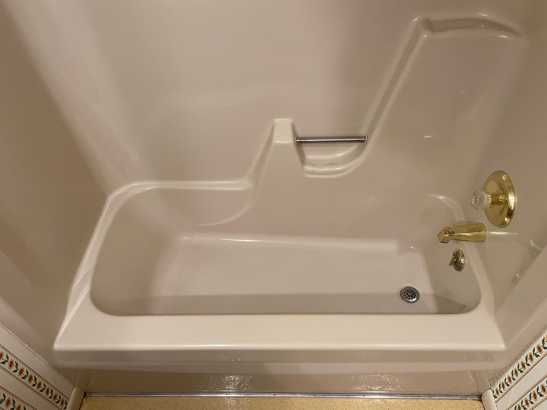 Tan walk-in bathtub with a built-in seat and a silver grab bar. The tub is inside a light-colored shower stall.