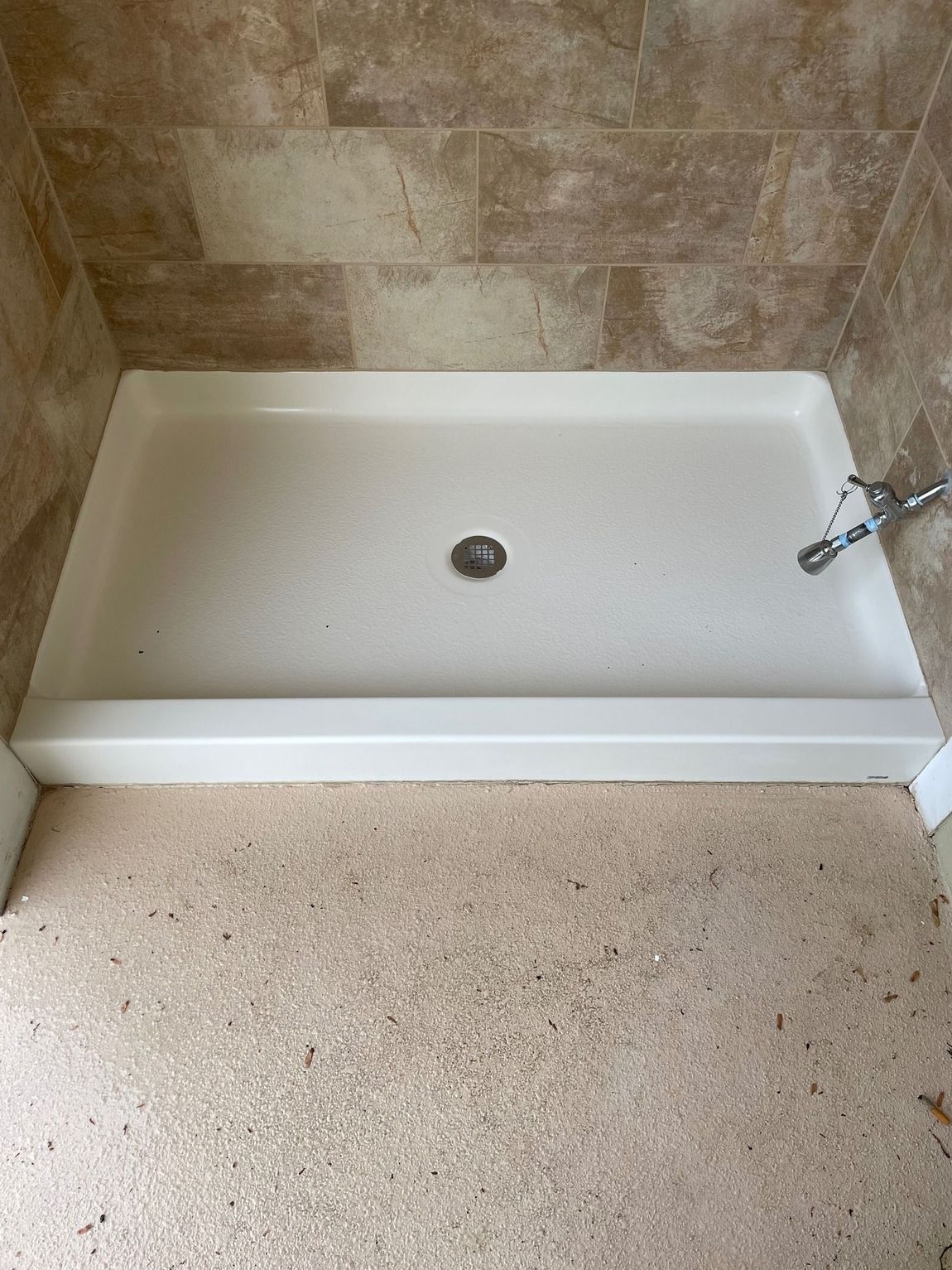 A shower stall with a white base and beige tile walls. The floor is speckled, and there's a faucet.