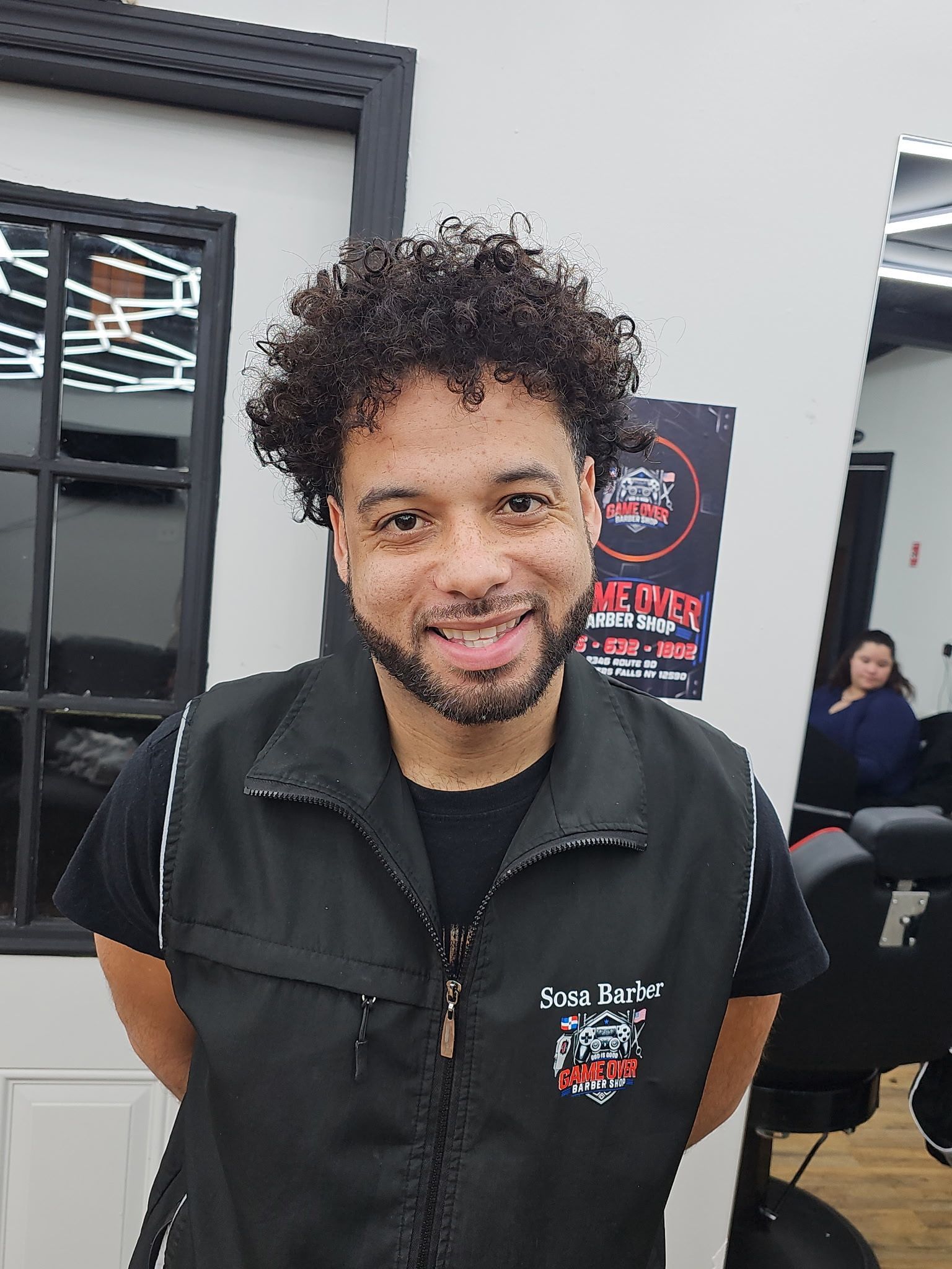 Smiling person with curly hair wearing a black vest in a salon with a mirror and chair in the background.