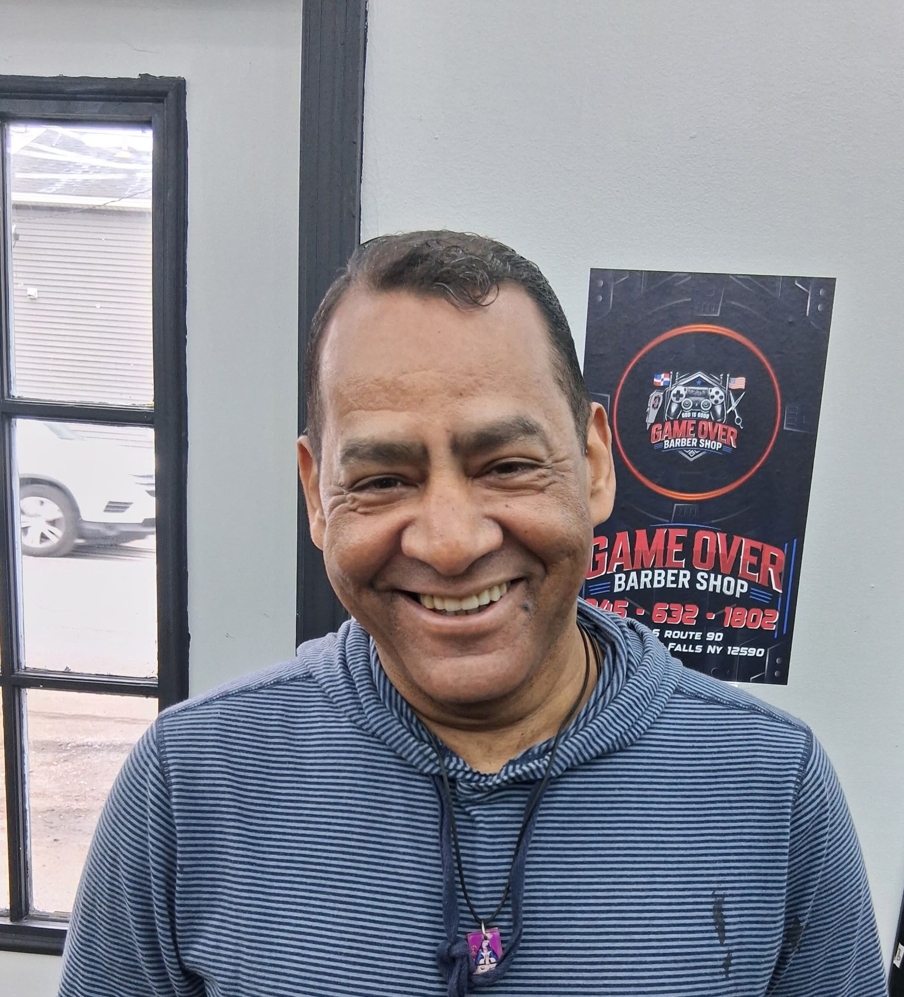 A smiling person wearing a blue striped hoodie, standing in front of a Game Over Barbershop sign.