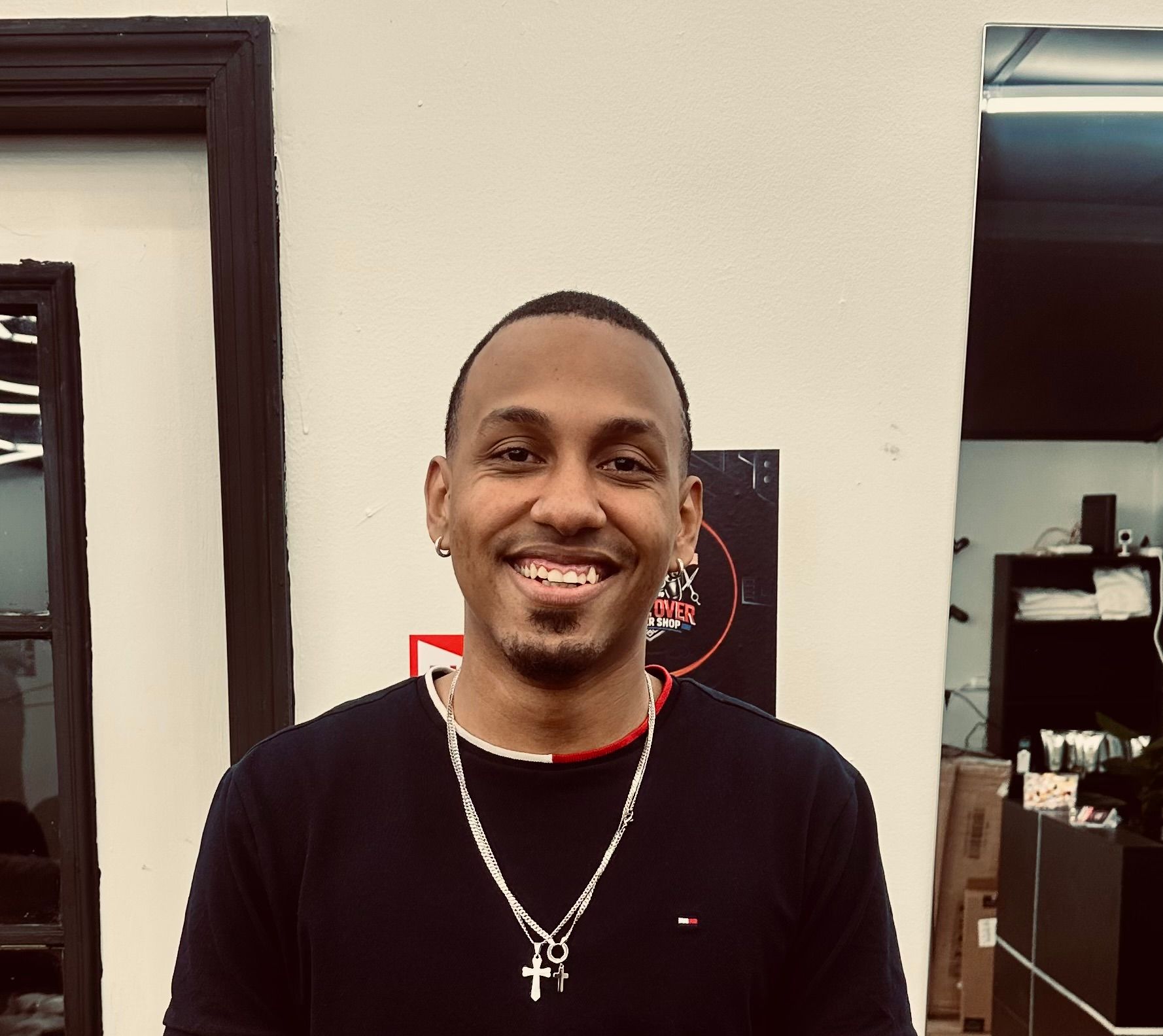 A person smiling while wearing a dark shirt and a silver chain with a cross pendant, standing in an indoor setting.