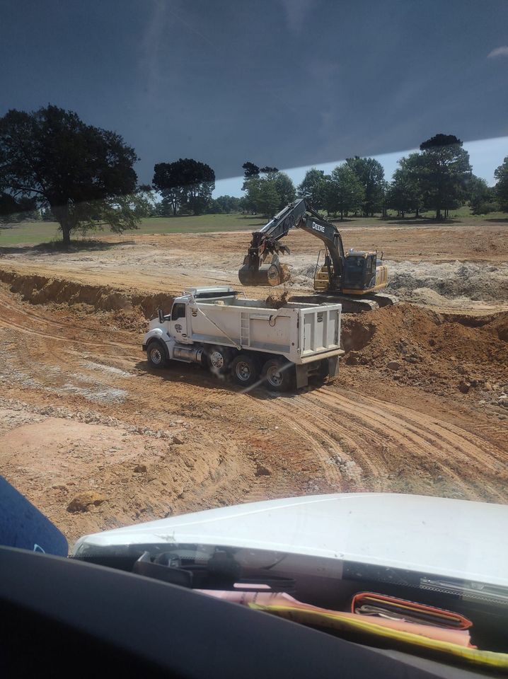 A dump truck is driving through a dirt field with an excavator.