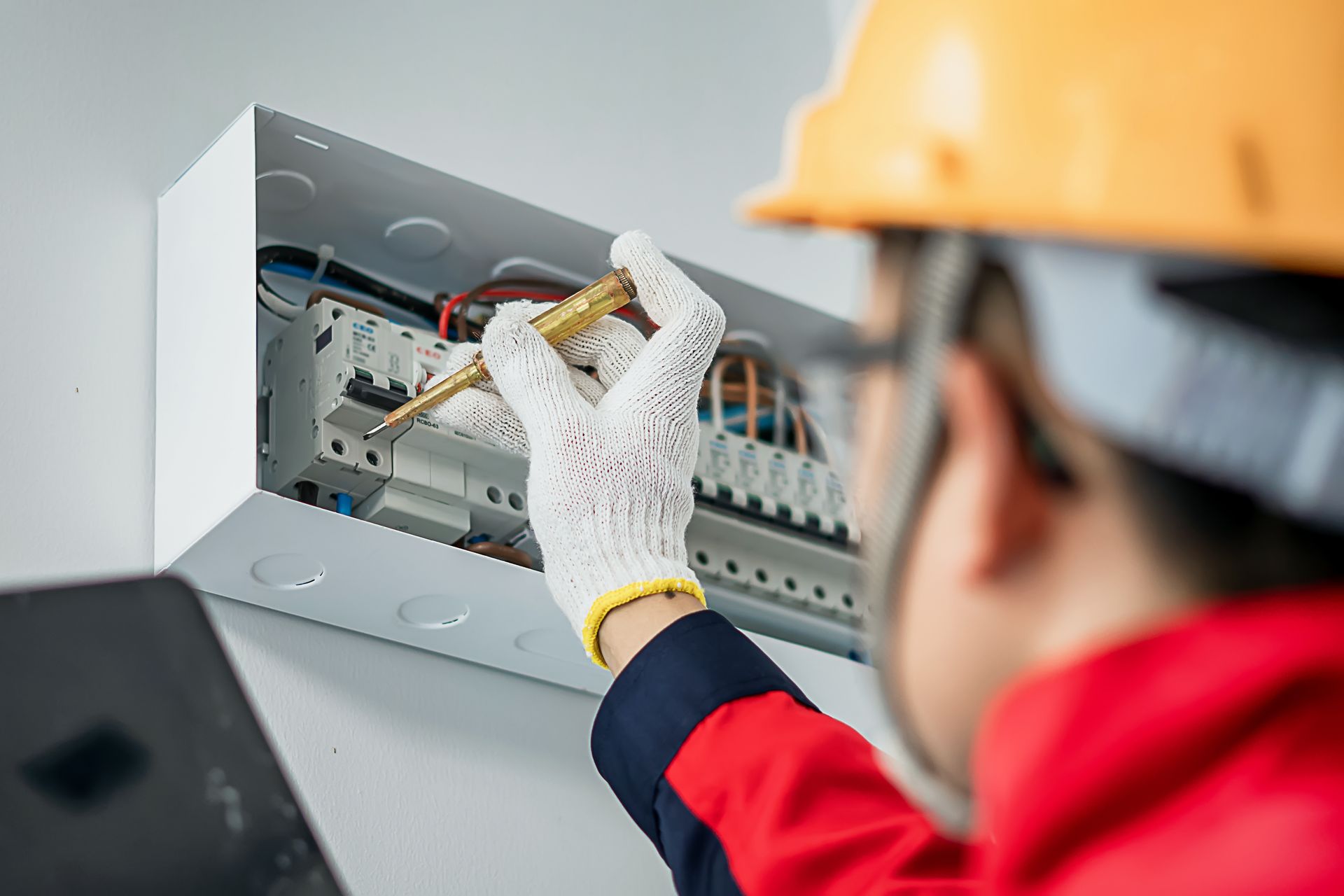 A man wearing a hard hat and gloves is working on an electrical box.