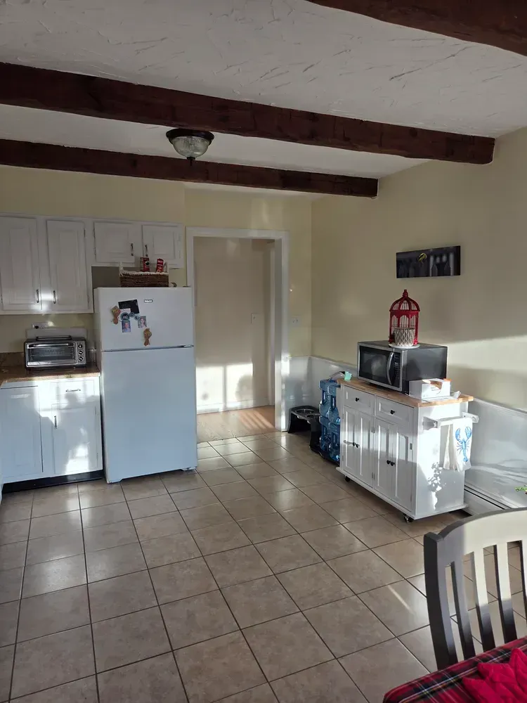Small kitchen with white cabinets, fridge, tiled floor, exposed beams, and a dining chair in the foreground