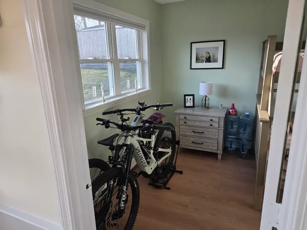Small room with two bicycles, a dresser, framed art, and water bottles by a window.
