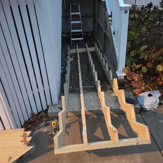 Wooden stair framing under construction beside a house, with ladder and tools in a narrow side yard