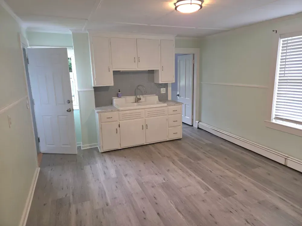 Empty apartment room with white cabinets, gray wood floors, and a window with blinds