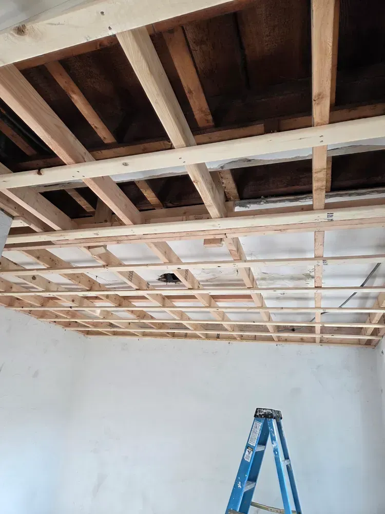 Partially finished room ceiling with exposed wooden framing and a blue step ladder below