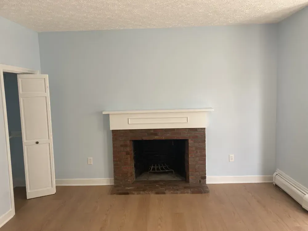 Empty living room with light blue walls, brick fireplace, white mantel, and wood floor.